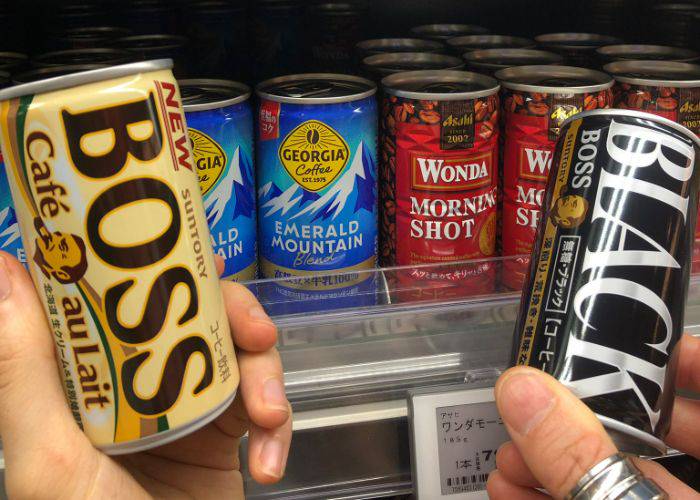 Two hands holding up Boss canned coffee in front of a fridge filled with different canned coffees.